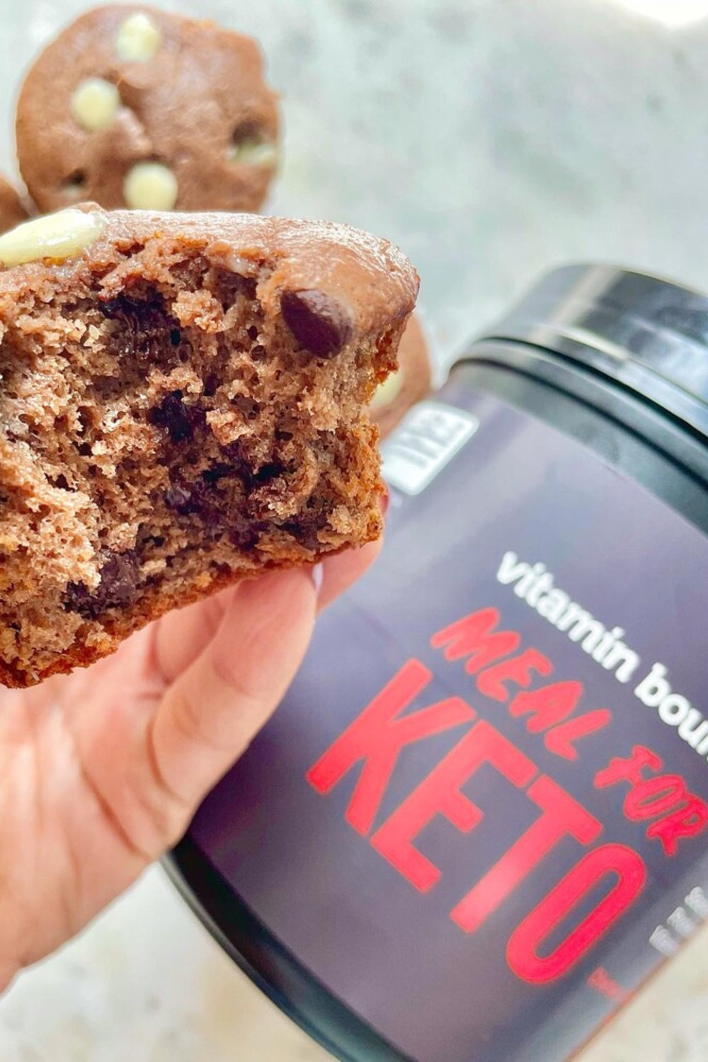 Meal for Keto Double Chocolate Chip Protein Muffins, pictured with a bite out of one, and Meal for Keto in the background